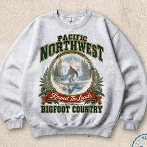 May include: A light gray crewneck sweatshirt featuring a graphic print with the text PACIFIC NORTHWEST, Respect The Locals, and BIGFOOT COUNTRY. The design includes a circular illustration of a Bigfoot figure in a snowy forest landscape.