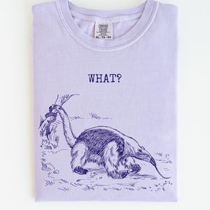 May include: A lavender Comfort Colors t-shirt with a navy blue illustration of an anteater and the word "WHAT?" printed above it. The shirt is folded and has a tag in the upper right corner.