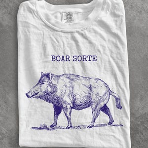 May include: White t-shirt with a blue illustration of a boar and the text "BOAR SORTE" above the image. The boar is drawn in a detailed, sketch-like style. The shirt is folded.