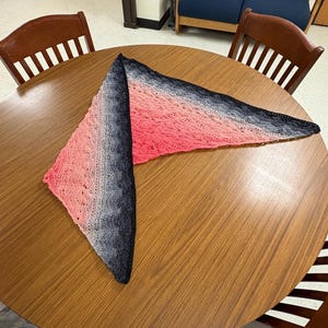 May include: A triangular crocheted shawl with a black, grey, and pink ombre pattern. The shawl is laid out on a wooden table.
