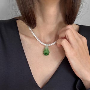 Könnte beinhalten: Eine Perlenkette mit einem grünen Anhänger. Der Anhänger hat eine einzigartige Form und eine strukturierte Oberfläche. Die Kette wird über einem schwarzen Oberteil getragen.