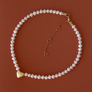 Puede incluir: Un delicado collar de perlas con un dije de corazón de oro. El collar está hecho de pequeñas perlas blancas y tiene un cierre de oro.