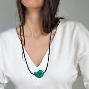 Pode incluir: Um colar longo com um pingente de concha turquesa e uma corrente de contas pretas. O pingente é de uma cor turquesa vibrante, e a corrente é composta por pequenas contas escuras. O colar é usado sobre uma blusa branca.