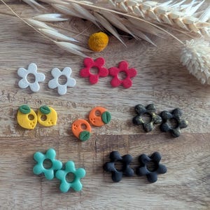 Pode incluir: Uma coleção de brincos artesanais de argila polimérica em vários designs florais e de frutas. Os brincos são brancos, vermelhos, amarelos, laranja, pretos e verde-azulado. Estão dispostos numa superfície de madeira com trigo seco e um pompom amarelo.