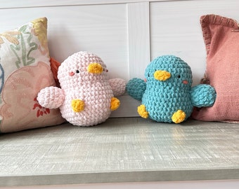 May include: Two crocheted bird plushies, one pink and one teal blue, with yellow beaks and feet. The plushies are sitting on a light grey wooden surface.