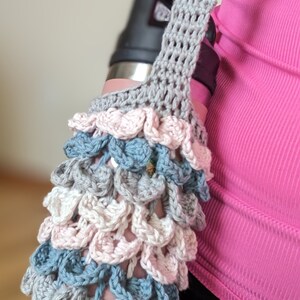 May include: A gray crocheted water bottle holder with a pink, white, and blue crocheted design. The holder is being worn by a person.