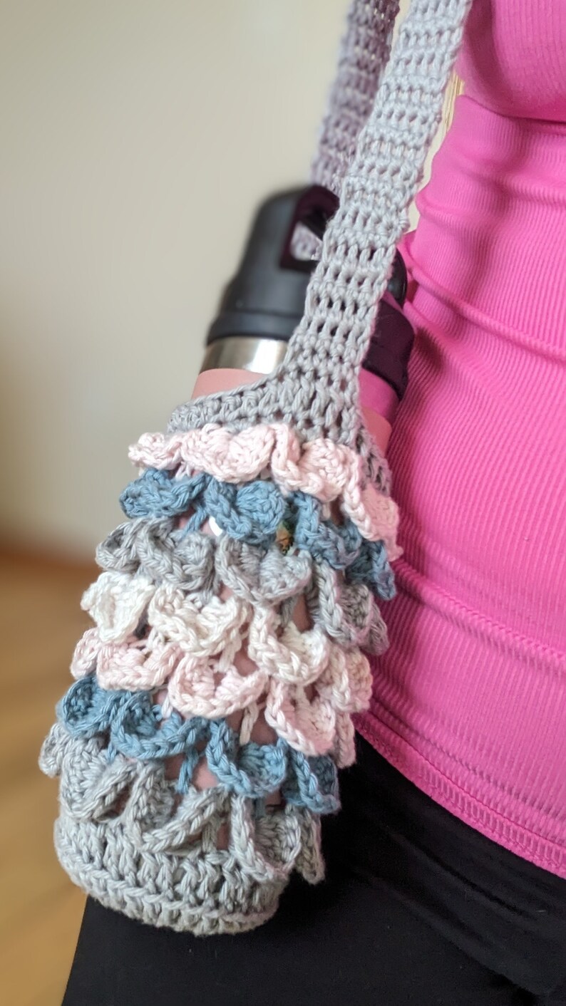 May include: A gray crocheted water bottle holder with a pink, white, and blue crocheted design. The holder is being worn by a person.