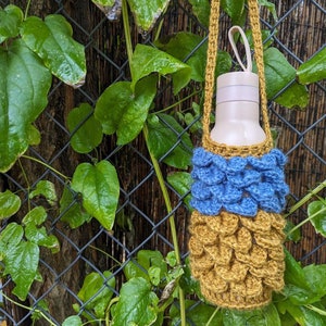 May include: A crocheted water bottle holder with a blue and gold pattern. The holder is hanging from a vine on a chain link fence.