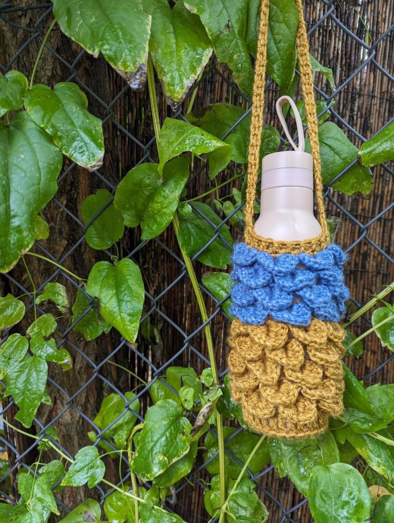 May include: A crocheted water bottle holder with a blue and gold pattern. The holder is hanging from a vine on a chain link fence.