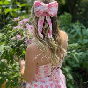 Op de afbeelding: Een roze en witte gingham gehaakte jurk met een bijpassende roze en witte gingham strik in het haar.