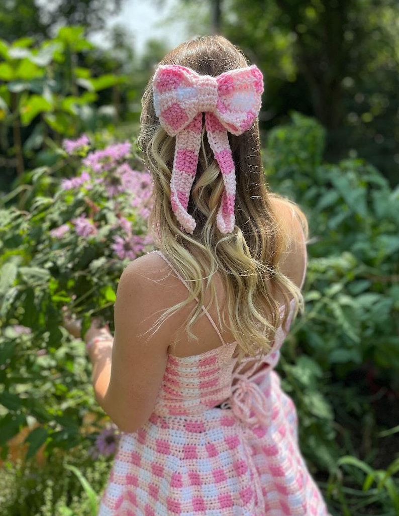 Op de afbeelding: Een roze en witte gingham gehaakte jurk met een bijpassende roze en witte gingham strik in het haar.