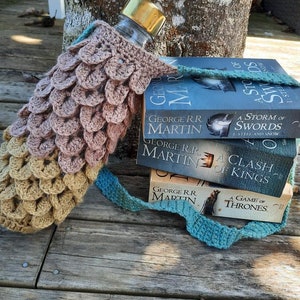 May include: A crocheted water bottle holder with a brown and beige pattern, holding a clear water bottle with a gold lid. The holder is wrapped around a stack of three books, all by George R.R. Martin, titled "A Storm of Swords", "A Clash of Kings", and "A Game of Thrones".