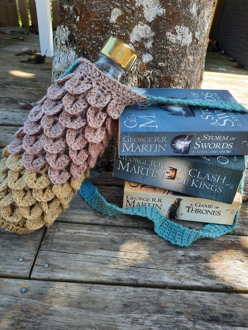 May include: A crocheted water bottle holder with a brown and beige pattern, holding a clear water bottle with a gold lid. The holder is wrapped around a stack of three books, all by George R.R. Martin, titled "A Storm of Swords", "A Clash of Kings", and "A Game of Thrones".