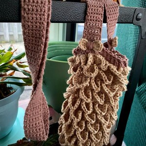 May include: Two crocheted bags hanging on a shelf. One bag is brown and the other is a light brown with a textured, scale-like pattern.