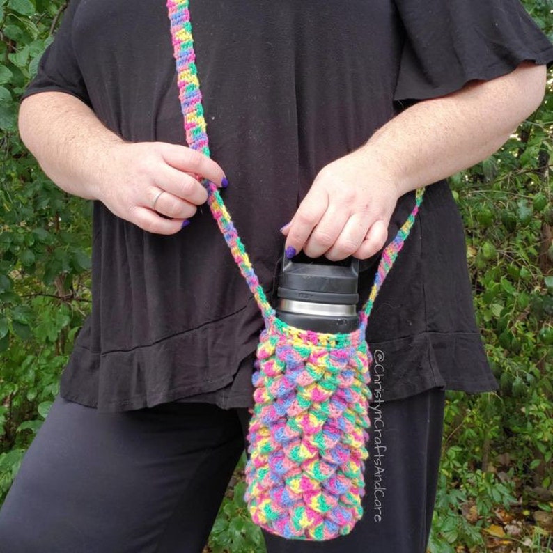 May include: A colorful crocheted water bottle holder with a shoulder strap. The holder is made with a scale pattern in pink, yellow, green, and blue. The holder is being worn by a person and is holding a stainless steel water bottle.