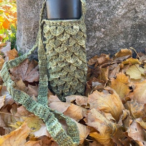 May include: A green crocheted bottle holder with a black lidded bottle inside. The bottle holder is lying on a bed of brown autumn leaves.