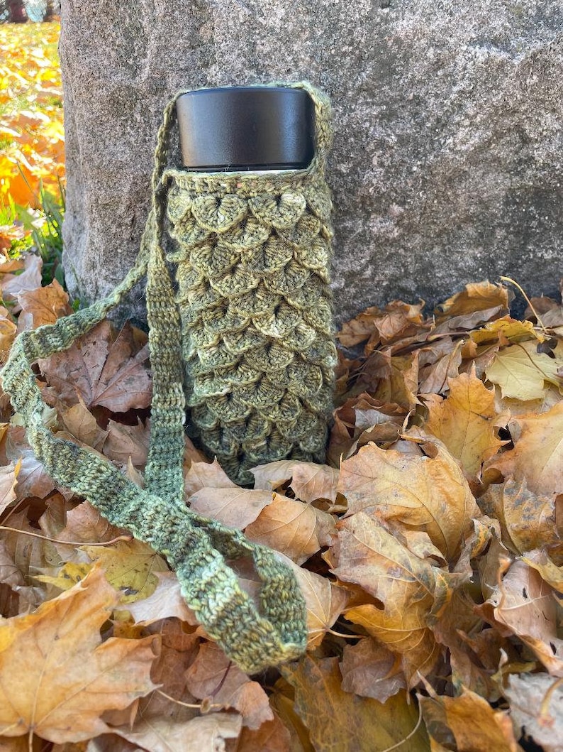 May include: A green crocheted bottle holder with a black lidded bottle inside. The bottle holder is lying on a bed of brown autumn leaves.