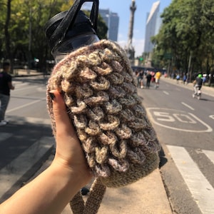 May include: A brown and beige crocheted water bottle holder with a strap. The holder is made with a textured, woven pattern.