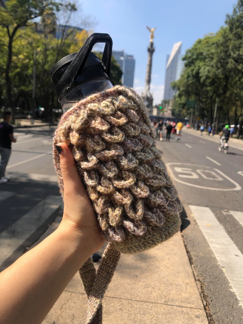 May include: A brown and beige crocheted water bottle holder with a strap. The holder is made with a textured, woven pattern.