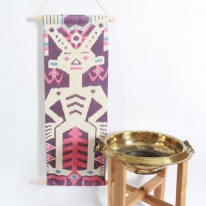 May include: A rectangular wall hanging with a geometric design in purple, pink, and cream colors. The hanging is suspended from a wooden rod. A brass bowl sits on a wooden stand.
