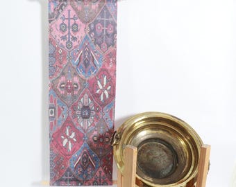 May include: A patterned wall hanging in pink, blue, and grey, suspended from a wooden rod. The design features geometric shapes. A large brass bowl sits on a wooden stand. The bowl is antique.