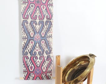 May include: A patterned textile hanging, featuring a geometric design in red, blue, and cream, suspended from a wooden rod. A large, oval brass tray sits on a wooden stand.