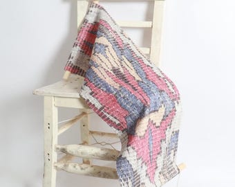 May include: A patterned textile in red, blue, beige, and grey hangs over a distressed white wooden chair. The textile, possibly a rug or wall hanging, features a geometric design. The chair adds a rustic touch to the scene, suggesting a bohemian aesthetic.