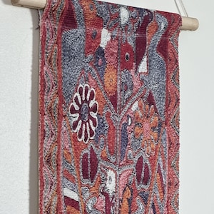 May include: A rectangular wall hanging with a detailed, colorful pattern. The design features a mix of red, orange, gray, and white elements, including floral and geometric shapes. It is suspended from a wooden dowel with a natural cord.
