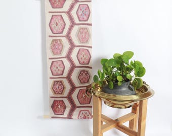May include: A patterned wall hanging featuring repeating hexagonal designs in pink, red, and beige tones. The textile is hung from a wooden rod. A small plant in a brass planter sits on a wooden stand.