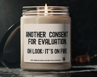 May include: A beige candle with a black label that reads "Another Consent for Evaluation. Oh Look: It's on Fire." The candle is lit and the flame is visible. The candle is on a dark surface.