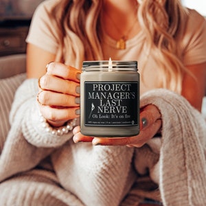 May include: A gray candle with a black label that reads "Project Manager's Last Nerve. Oh Look: It's on fire." The candle is lit and being held by a person.