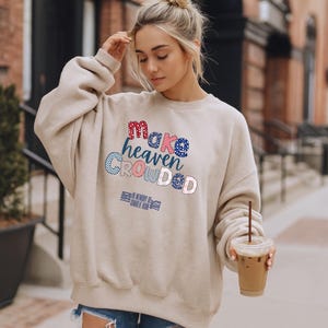 May include: Beige sweatshirt with the text "Make Heaven Crowded" in red, blue, and pink lettering. The sweatshirt also has the text "1953 In Memory Of Charlie Kay" in blue. The sweatshirt is a casual style.