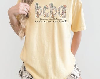 May include: A light yellow t-shirt with the text "bcba" in a floral design and the words "board certified behavior analyst" in black script. The shirt is a short-sleeved, casual style.