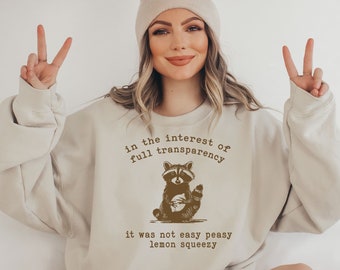 May include: A beige sweatshirt with a brown graphic of a raccoon holding a lemon. The text on the sweatshirt reads "in the interest of full transparency it was not easy peasy lemon squeezy."