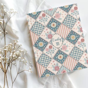 Peut inclure: Un journal à motifs avec un design matelassé. La couverture présente un mélange de couleurs rose, bleu et crème avec des accents de marguerites et de fleurs. Le journal porte le nom "Emily" et l'année "2026" imprimés sur la couverture.