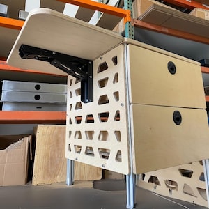 Może przedstawiać: Jasne drewniane biurko ze składanym blatem i dwiema szufladami. Biurko ma unikalny design z wyciętymi wzorami po bokach i jest wspierane przez metalowe nogi. Blat jest przymocowany czarnymi metalowymi zawiasami.