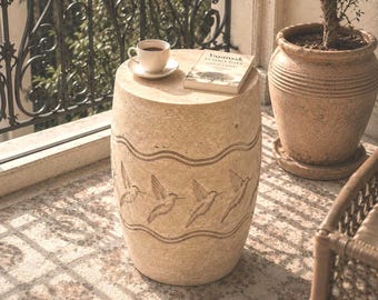 Puede incluir: Un taburete de jardín cilíndrico de piedra beige con una banda horizontal tallada que muestra cuatro colibríes en vuelo entre dos líneas onduladas. Una taza de café blanca sobre un plato y un libro titulado Yaşamak Cesaret İster descansan encima.