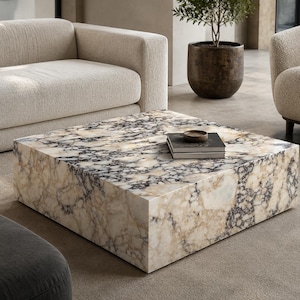 Puede incluir: Una mesa de centro cuadrada con un patrón similar al mármol en tonos beige, crema y gris oscuro. La mesa es el punto focal de una sala de estar moderna, con un sofá y un sillón en el fondo. Una pequeña pila de libros y un cuenco encima.