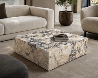 Puede incluir: Una mesa de centro cuadrada con un patrón similar al mármol en tonos beige, crema y gris oscuro. La mesa es el punto focal de una sala de estar moderna, con un sofá y un sillón en el fondo. Una pequeña pila de libros y un cuenco encima.