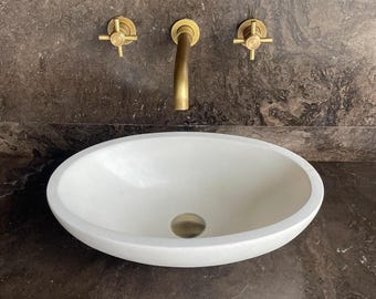 Puede incluir: Un lavabo ovalado blanco con un desagüe y un grifo de latón. Dos manijas de latón están montadas en la pared sobre el lavabo. El lavabo está colocado contra una pared oscura y veteada.
