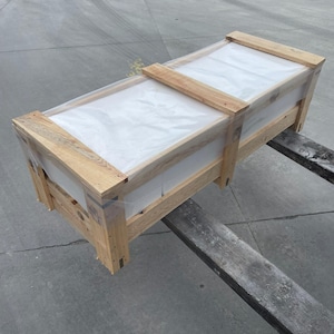May include: A rectangular wooden crate with a clear plastic top, secured with wooden slats. The crate is on a forklift, ready for transport. The wood has a natural, light brown colour.