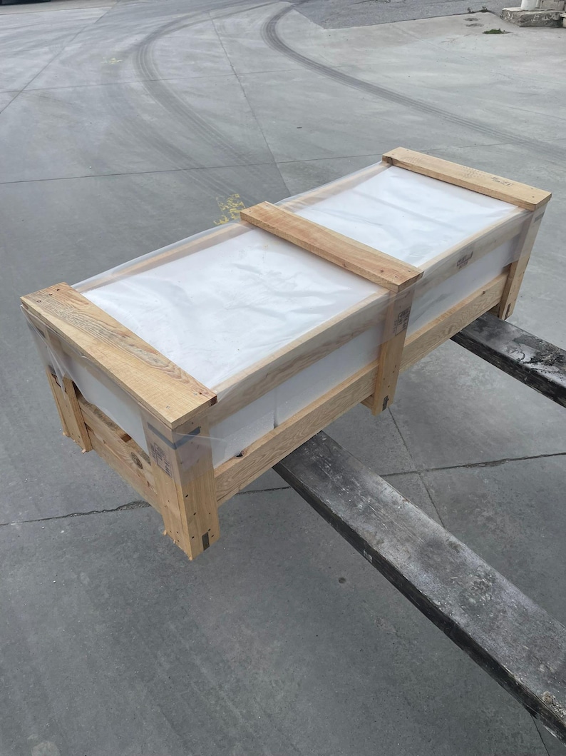 May include: A rectangular wooden crate with a clear plastic top, secured with wooden slats. The crate is on a forklift, ready for transport. The wood has a natural, light brown colour.