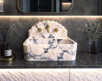 Puede incluir: Un lavabo de baño de mármol con un patrón veteado blanco y gris. El lavabo tiene un protector contra salpicaduras festoneado y grifos dorados. El lavabo está colocado contra una pared gris oscuro con un espejo encima.
