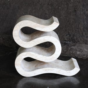 Puede incluir: Una mesa auxiliar escultural en forma de serpentina, de color crema. La mesa tiene un dise&ntilde;o liso y curvado con tres niveles distintos. La pieza est&aacute; hecha de un material s&oacute;lido y claro y se apoya sobre una superficie oscura y reflectante.