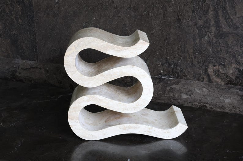 Puede incluir: Una mesa auxiliar escultural en forma de serpentina, de color crema. La mesa tiene un dise&ntilde;o liso y curvado con tres niveles distintos. La pieza est&aacute; hecha de un material s&oacute;lido y claro y se apoya sobre una superficie oscura y reflectante.