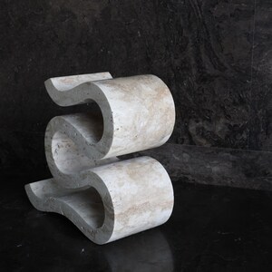 Puede incluir: Una escultura abstracta &uacute;nica, de color beige claro, con un dise&ntilde;o ondulado en forma de S. La escultura est&aacute; hecha de un material similar a la piedra y se encuentra sobre una superficie oscura y reflectante. Tiene una est&eacute;tica moderna y art&iacute;stica.