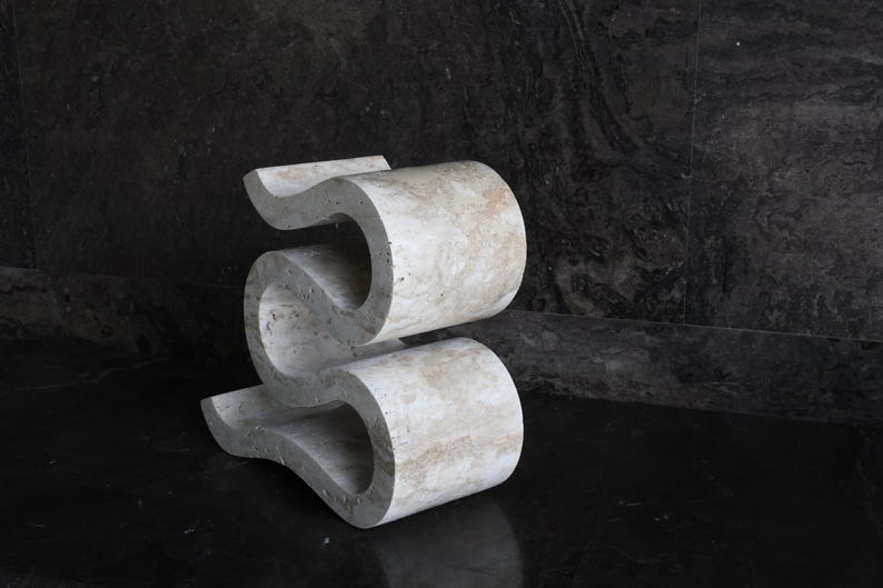 Puede incluir: Una escultura abstracta &uacute;nica, de color beige claro, con un dise&ntilde;o ondulado en forma de S. La escultura est&aacute; hecha de un material similar a la piedra y se encuentra sobre una superficie oscura y reflectante. Tiene una est&eacute;tica moderna y art&iacute;stica.