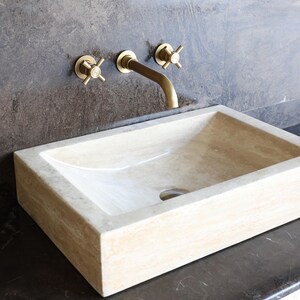 May include: A rectangular, beige stone sink with a curved interior sits on a dark countertop. Above the sink, a gold-colored faucet and two matching cross-handle valves are mounted on a dark gray wall. The sink has a drain hole.