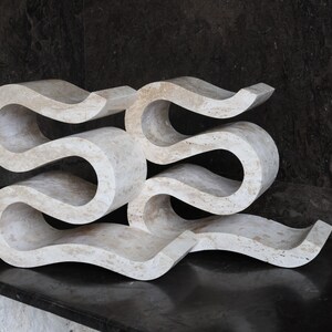 Puede incluir: Una escultura abstracta de color crema con un dise&ntilde;o fluido en forma de ola. La escultura est&aacute; hecha de un material s&oacute;lido y tiene un acabado liso y pulido. La pieza se exhibe sobre una superficie oscura, creando un contraste.