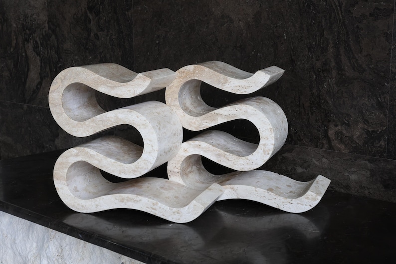 Puede incluir: Una escultura abstracta de color crema con un dise&ntilde;o fluido en forma de ola. La escultura est&aacute; hecha de un material s&oacute;lido y tiene un acabado liso y pulido. La pieza se exhibe sobre una superficie oscura, creando un contraste.